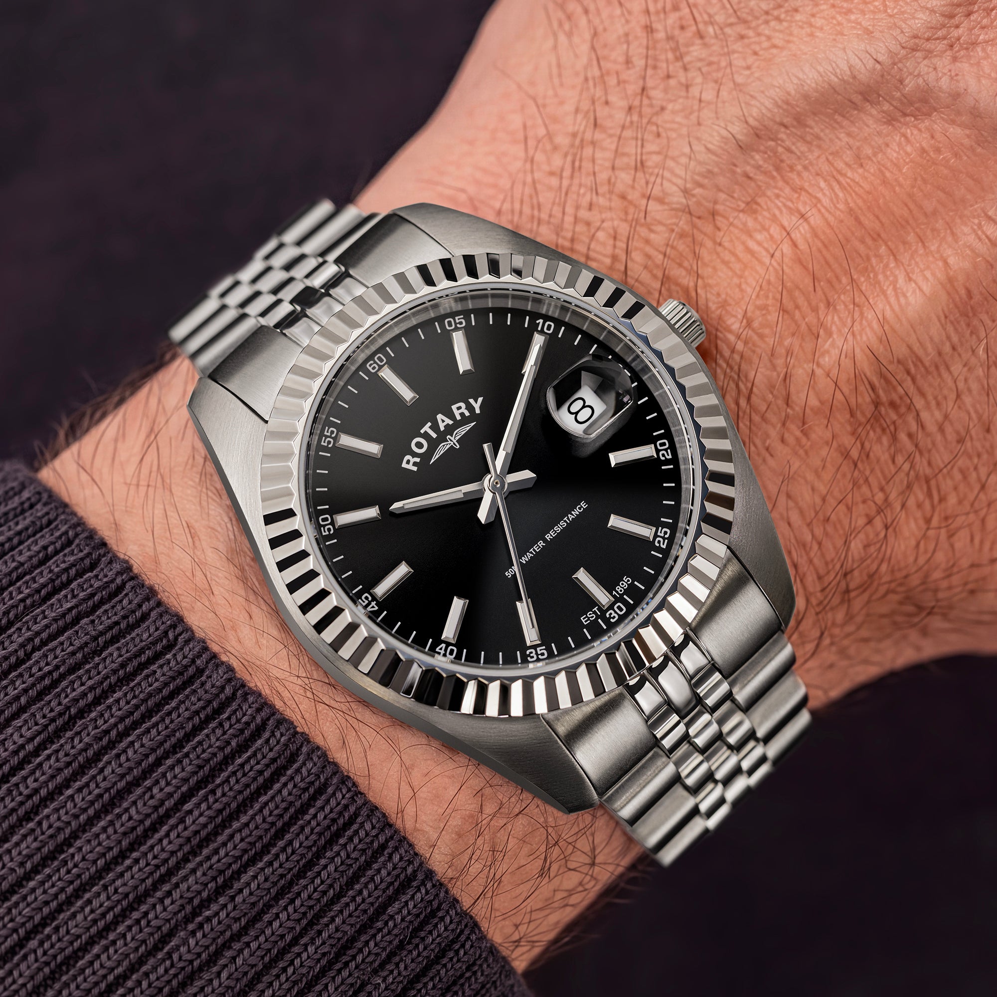 A close-up of a wrist wearing a Rotary stainless steel watch with a black dial, fluted bezel, and metal bracelet.