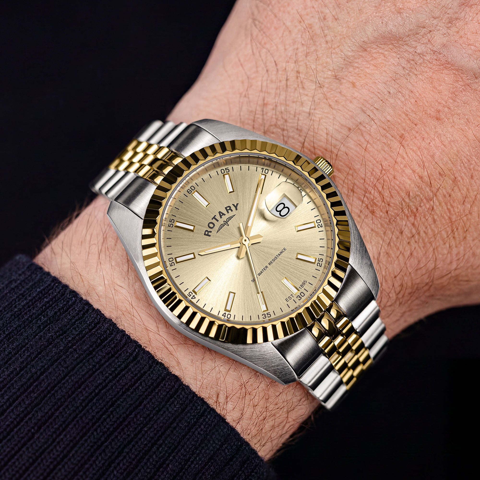 A two-tone Rotary stainless steel and gold wristwatch with a fluted bezel and champagne dial, shown worn on a person's wrist against a dark background.