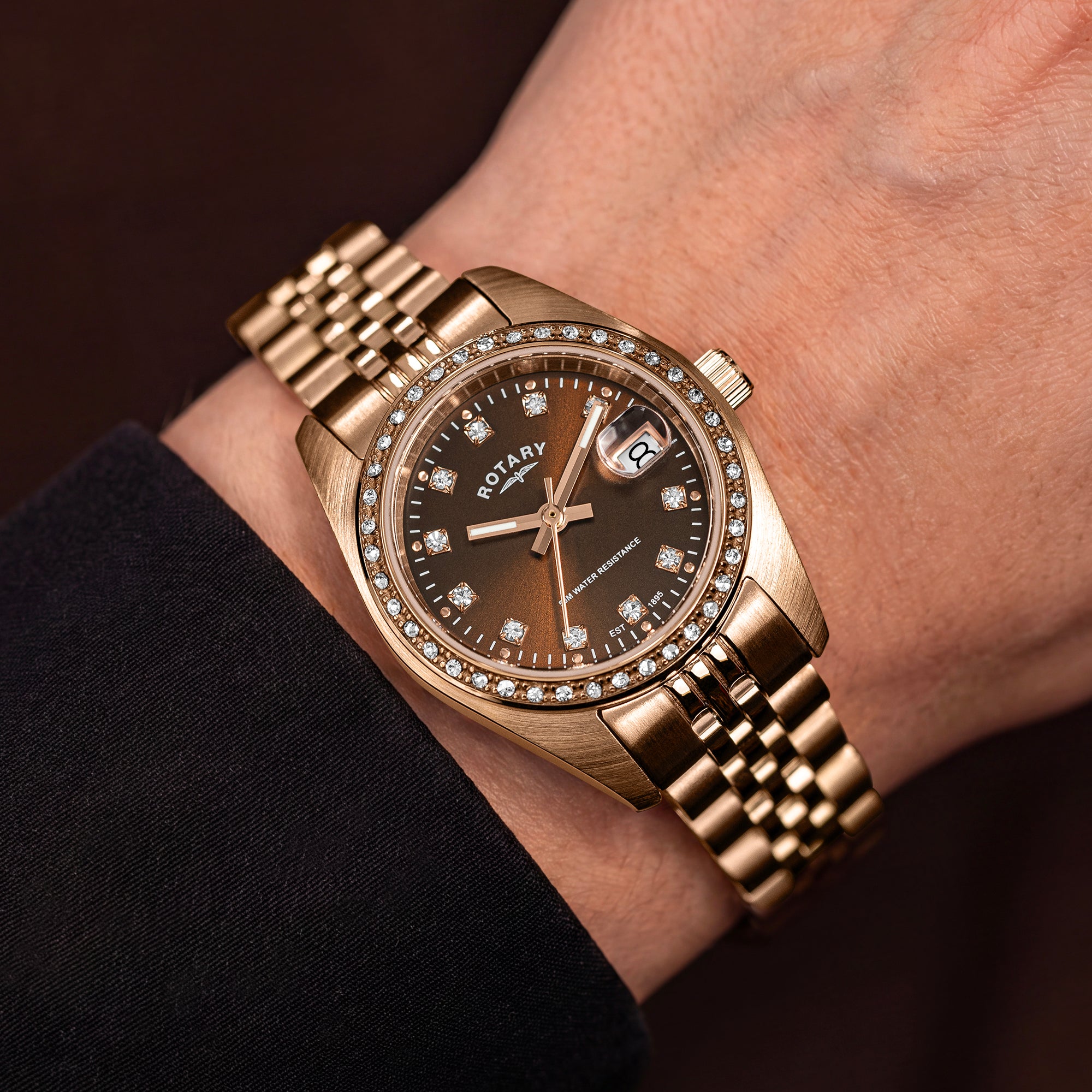 A close-up of a Rotary Dress Date watch with a rose gold-tone stainless steel bracelet and case, featuring a dark brown dial and a bezel set with small diamonds, worn on a wrist.
