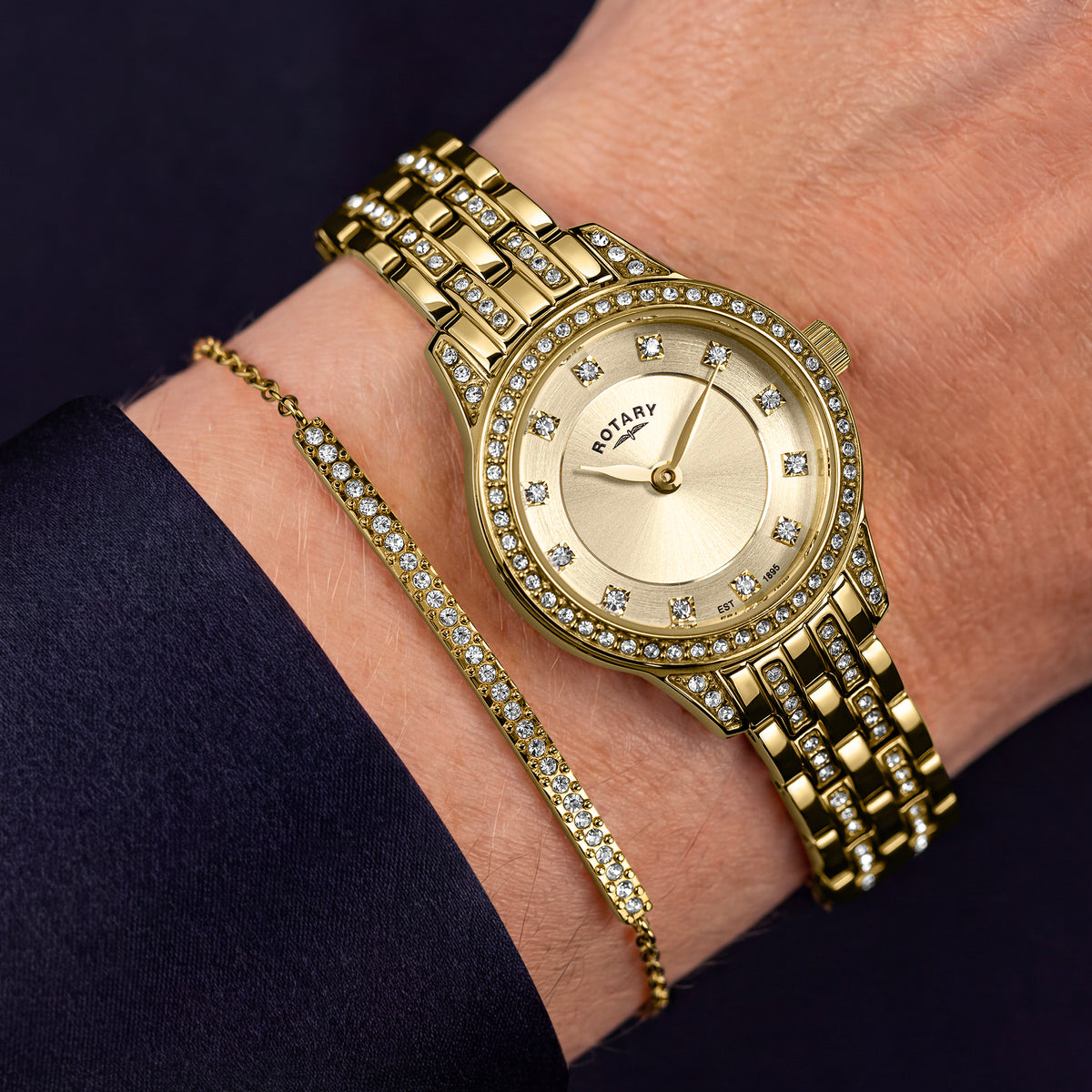 A gold-tone watch with a gem-set bezel and matching crystal-encrusted bracelet is shown on a person's wrist, alongside a slim matching crystal bracelet. The watch brand 'ROTARY' is visible on the dial.