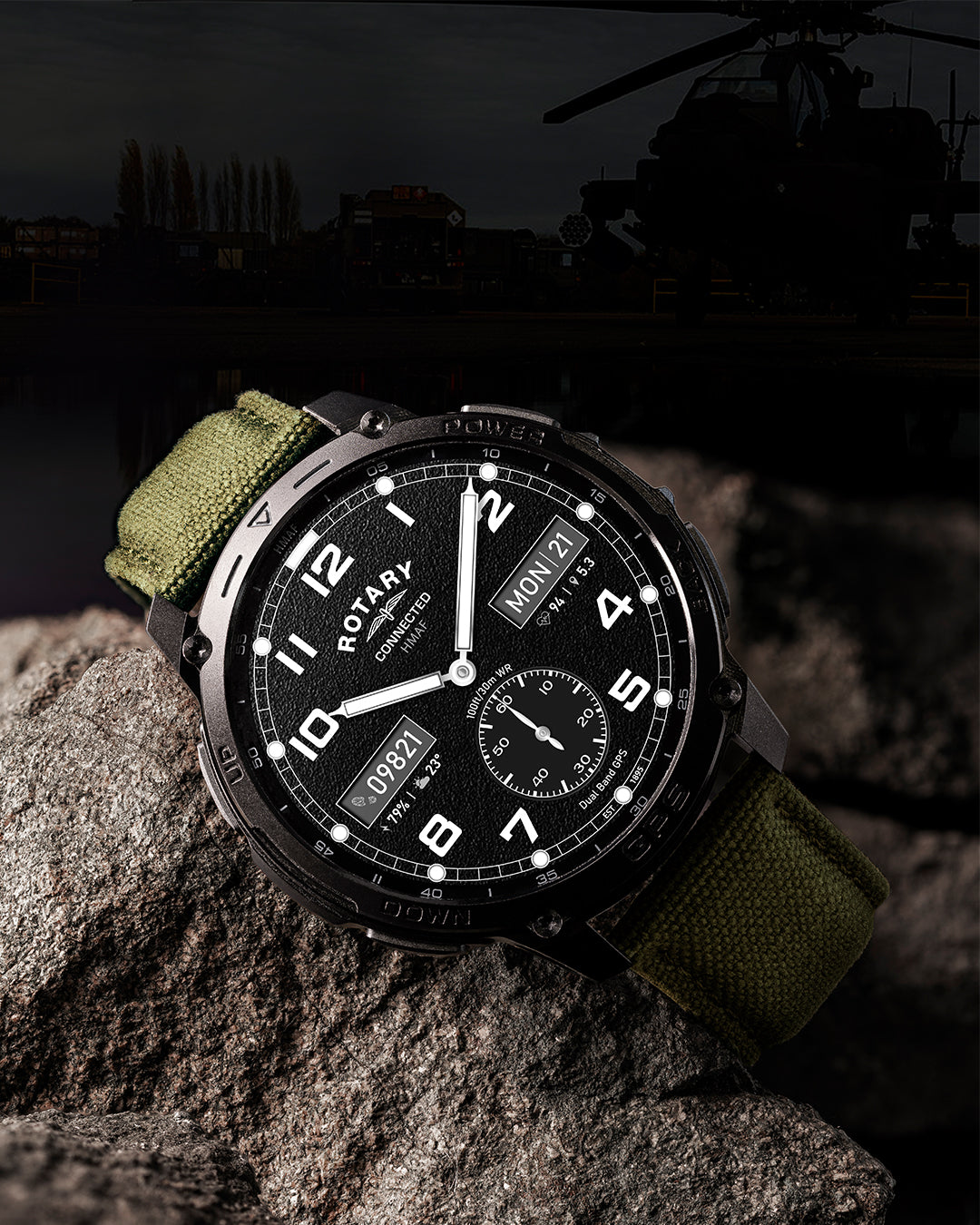 A Rotary x MoD Connected smartwatch with a black dial, silver-tone case, and olive green fabric strap, resting on a textured rock with a dark, moody outdoor background featuring a helicopter silhouette.