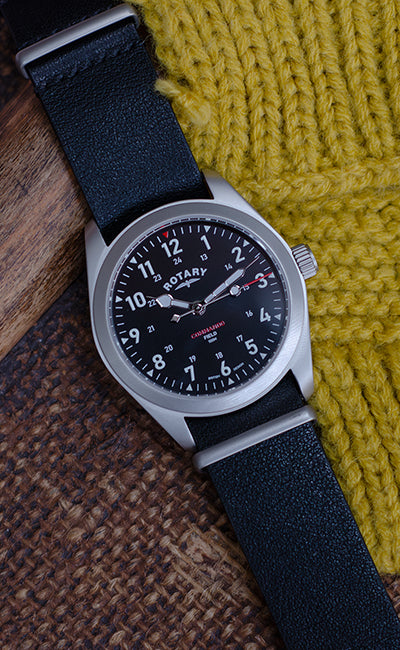 a rotary field watch in stainless steel with a black dial and black leather strap on a wooden table with a yellow cloth