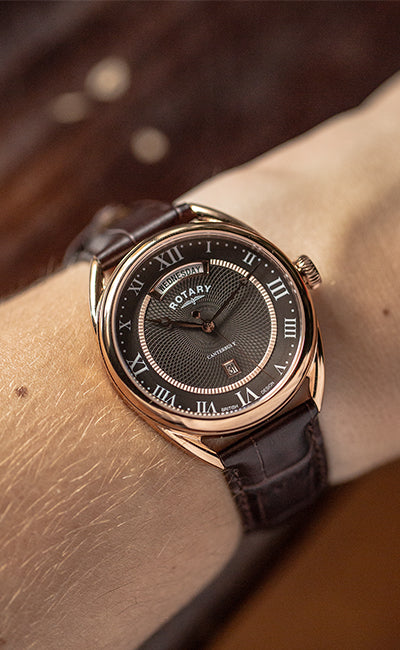 a man wearing a traditional style rotary watch in rose gold with a brown leather strap and black dial