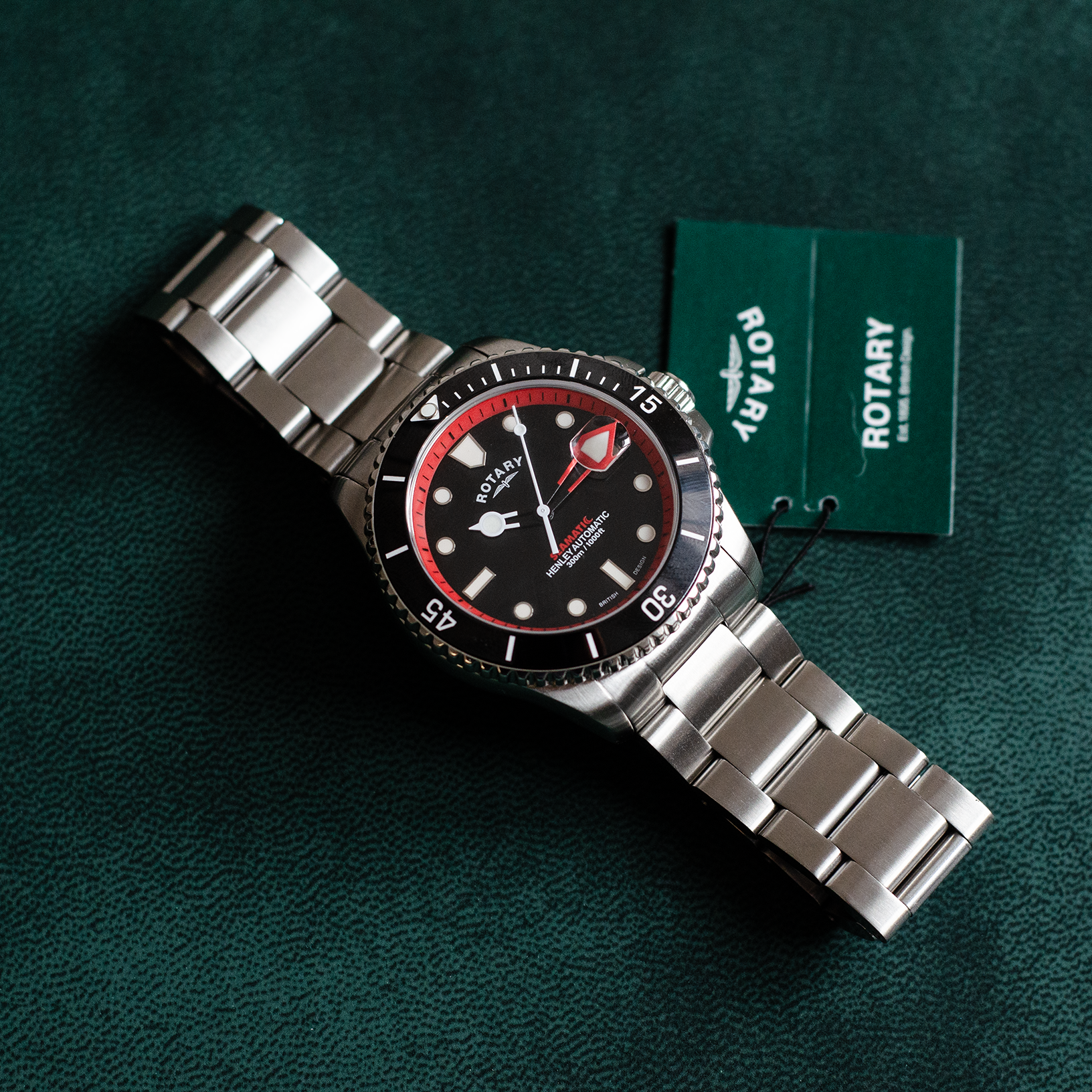 a Rotary seamatic dive watch in stainless steel lying on a green leather mat with a Rotary swing tag attached to the strap. The black dial has red coloured accenting and 3 white hands with a ceramic black bezel