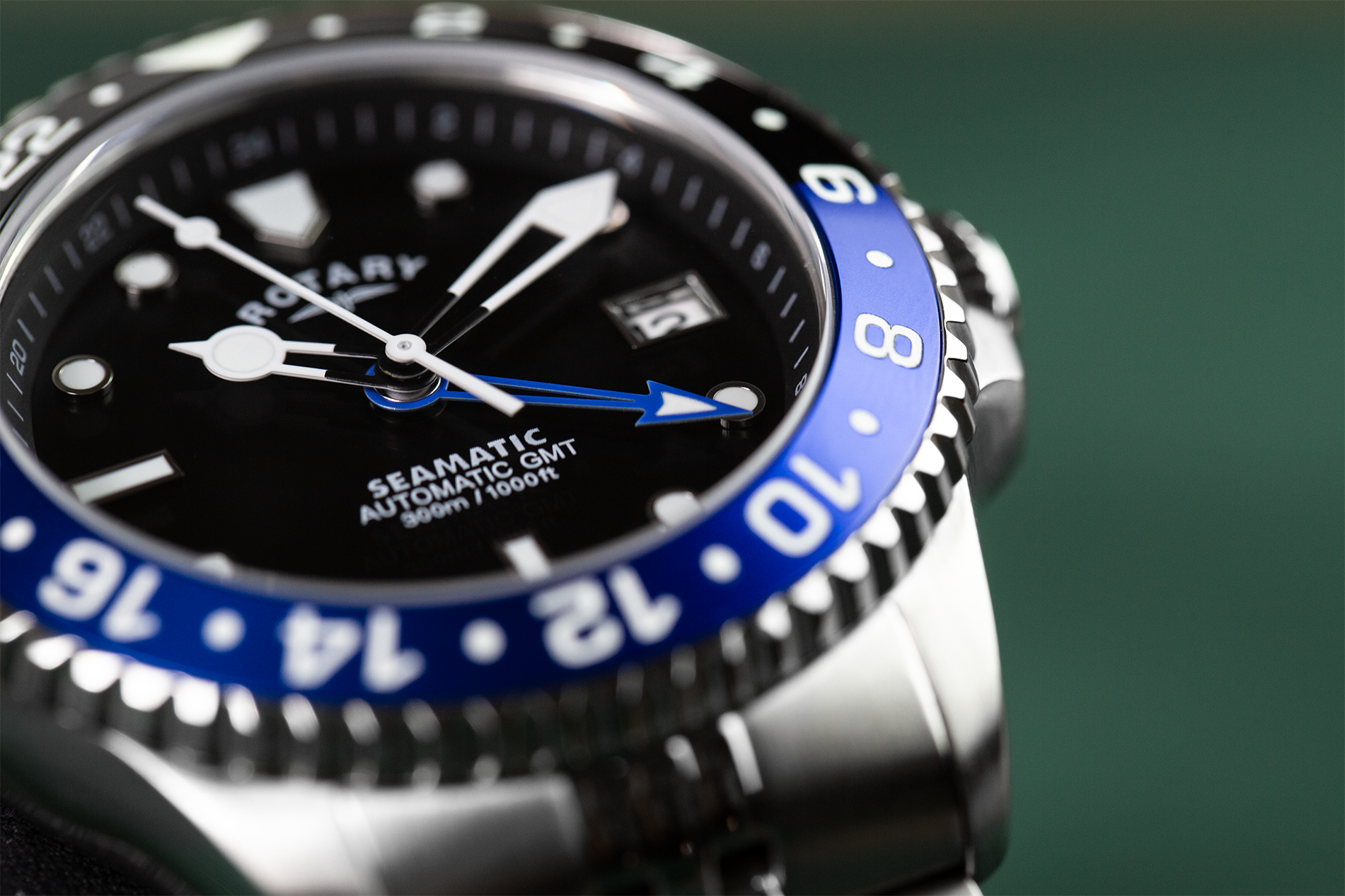 Close-up of a Rotary Seamatic 300 GMT Automatic watch with a black dial, blue bezel, and silver stainless steel case against a dark green background.