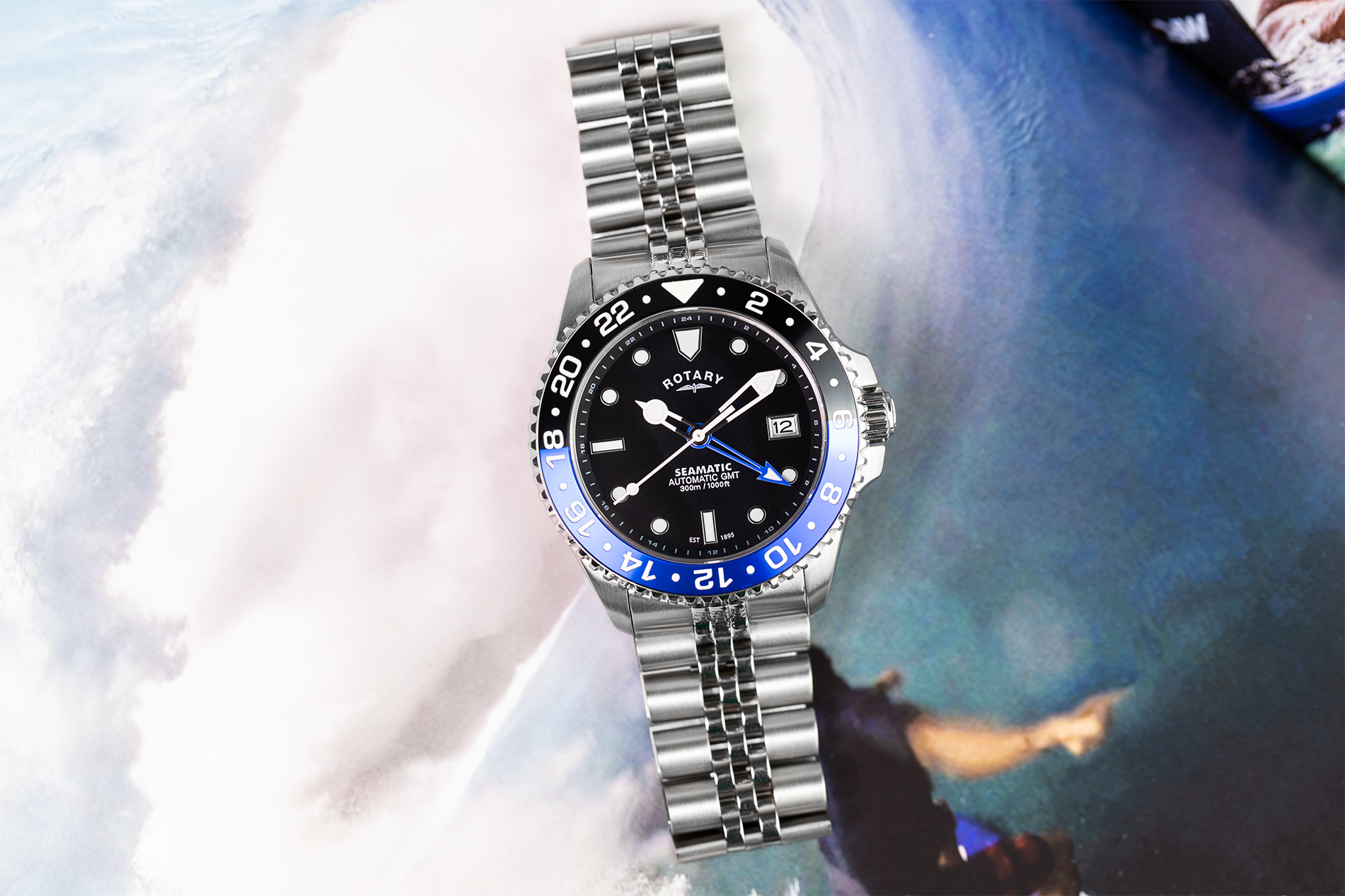 A stainless steel Rotary Seamatic 300 GMT wristwatch with a black dial, blue bezel, and silver link bracelet, shown against a blurred background of white and blue tones.